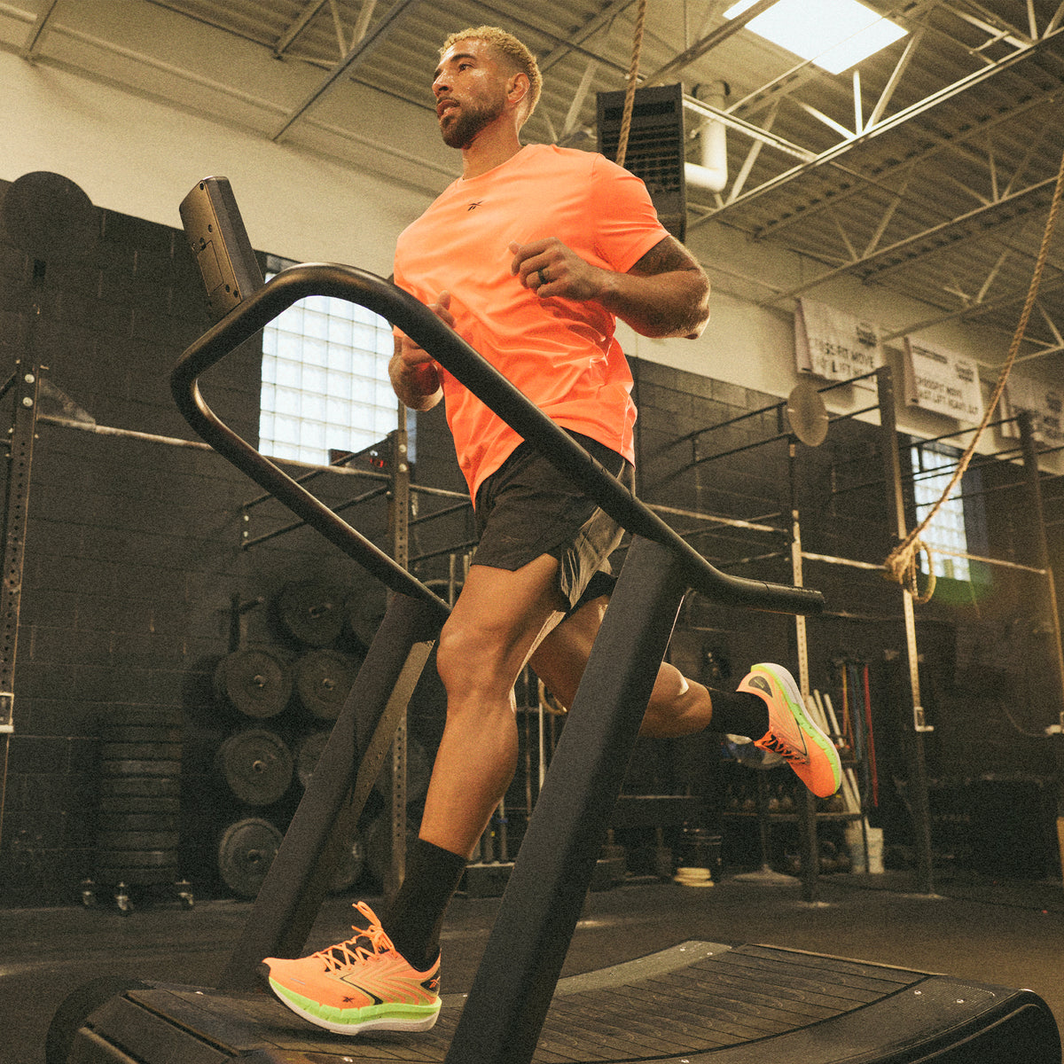 A man in a bright orange shirt and black shorts runs on a treadmill in a gym with black walls, weights, and workout equipment visible in the background.