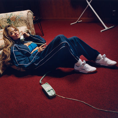 A woman in a blue tracksuit lies on a red carpet, talking on a green corded phone. She leans against a floral sofa, with an ironing board in the background. She wears white sneakers and looks relaxed.