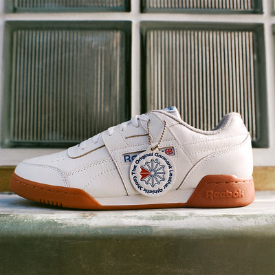 White Reebok sneaker with brown sole on a concrete surface with glass block wall background