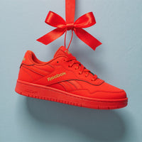 A bright red Reebok sneaker with gold logo lettering, hanging from a matching red ribbon bow against a light blue-gray background.